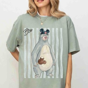 May include: A sage green short-sleeve t-shirt featuring a graphic of a grey bear wearing a black ear hat with a red heart icon. The bear holds a brown chocolate-covered treat. The text "Baloo" is written in black script above the bear.