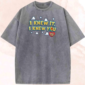 May include: A charcoal grey short-sleeve t-shirt featuring a graphic print with the text I KNEW IT, I KNEW YOU in yellow and blue letters. The design includes white cloud and star icons, along with a red heart containing the letters TS.
