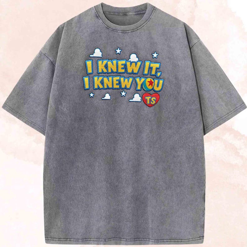 May include: A charcoal grey short-sleeve t-shirt featuring a graphic print with the text I KNEW IT, I KNEW YOU in yellow and blue letters. The design includes white cloud and star icons, along with a red heart containing the letters TS.