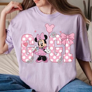 May include: A light purple t-shirt featuring the word "GIGI" in pink polka dot letters. Minnie Mouse is in the center, holding a microphone. A pink bow and balloon complete the design. The shirt is a relaxed fit.