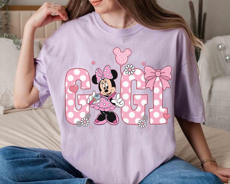 May include: A light purple t-shirt featuring the word "GIGI" in pink polka dot letters. Minnie Mouse is in the center, holding a microphone. A pink bow and balloon complete the design. The shirt is a relaxed fit.