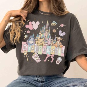 May include: Dark gray t-shirt featuring a colorful Disney World graphic. The design includes the Disney World castle, Mickey Mouse ears, and various Disney characters. The text "Disneyworld" is prominently displayed across the front.