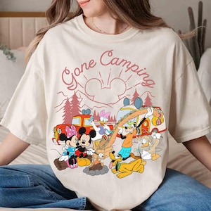 Pu&ograve; includere: T-shirt color crema con una grafica di personaggi Disney in campeggio. Il design include Topolino e Minnie, Paperino, Paperina, Pippo e Pluto attorno a un fal&ograve;, con la scritta "Gone Camping".