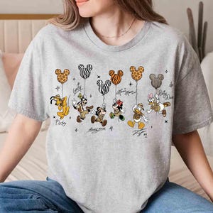 May include: A light gray t-shirt featuring Disney characters Pluto, Goofy, Mickey Mouse, Minnie Mouse, Donald Duck, and Daisy Duck. Each character is holding a Mickey Mouse-shaped balloon with a unique pattern. The characters' names and signatures are below them.