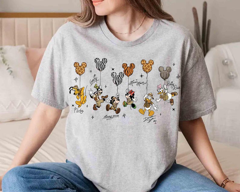 May include: A light gray t-shirt featuring Disney characters Pluto, Goofy, Mickey Mouse, Minnie Mouse, Donald Duck, and Daisy Duck. Each character is holding a Mickey Mouse-shaped balloon with a unique pattern. The characters' names and signatures are below them.