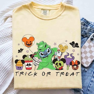 May include: A light yellow t-shirt featuring a Halloween-themed graphic with the text "TRICK OR TREAT". The design includes cartoon characters, bats, and Mickey Mouse-shaped balloons and treats. The shirt is folded on a white surface.