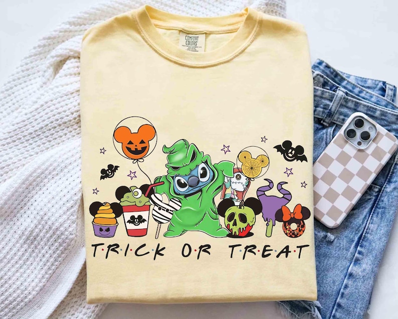 May include: A light yellow t-shirt featuring a Halloween-themed graphic with the text "TRICK OR TREAT". The design includes cartoon characters, bats, and Mickey Mouse-shaped balloons and treats. The shirt is folded on a white surface.