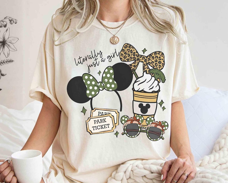 May include: Cream-colored t-shirt with a graphic design featuring a Minnie Mouse ear headband, sunglasses, a coffee cup, and a park ticket. The text "literally just a girl" is printed on the shirt.