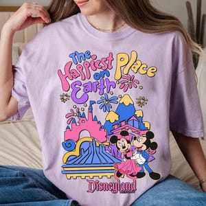 K&ouml;nnte beinhalten: Helllila T-Shirt mit farbenfroher Grafik. Das Design zeigt Micky und Minnie Maus vor einem Schloss und den Text "The Happiest Place on Earth" und "Disneyland Resort".