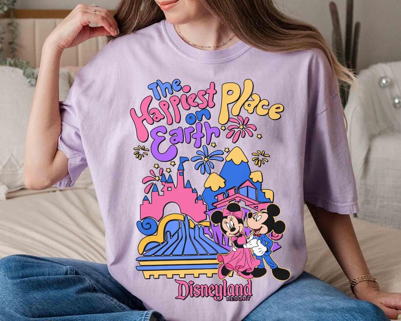 K&ouml;nnte beinhalten: Helllila T-Shirt mit farbenfroher Grafik. Das Design zeigt Micky und Minnie Maus vor einem Schloss und den Text "The Happiest Place on Earth" und "Disneyland Resort".