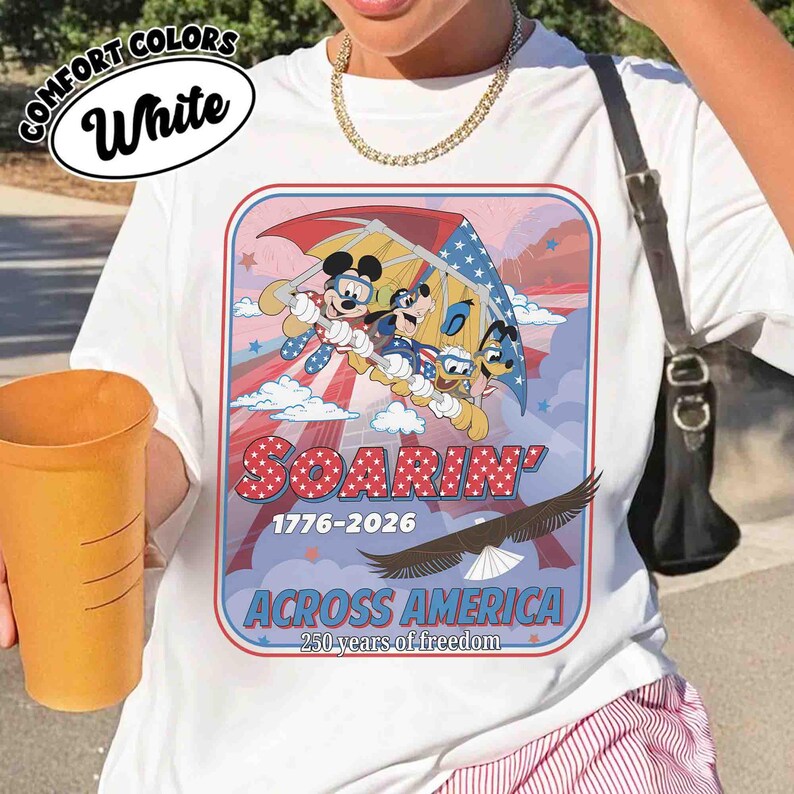 May include: White t-shirt with a graphic featuring Mickey Mouse, Goofy, and Donald Duck soaring with an American flag-themed parachute. The design includes the text "SOARIN' 1776-2026 ACROSS AMERICA 250 years of freedom."