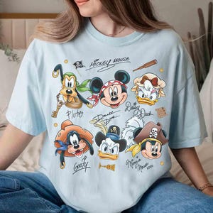 May include: Light blue t-shirt featuring a graphic of Disney characters as pirates, including Mickey Mouse, Donald Duck, and Goofy. The shirt has character signatures and the words "Mickey Mouse" at the top.