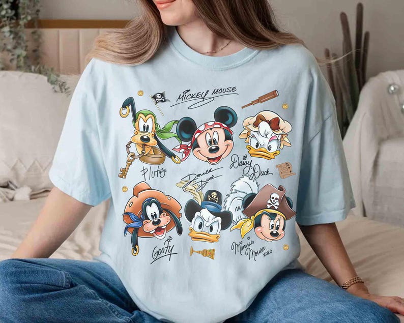 May include: Light blue t-shirt featuring a graphic of Disney characters as pirates, including Mickey Mouse, Donald Duck, and Goofy. The shirt has character signatures and the words "Mickey Mouse" at the top.