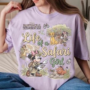 May include: A light purple t-shirt featuring a graphic design with the text "Life of a Safari Girl" and Disney's Animal Kingdom logo. The design includes illustrations of Simba, Timon, Pumbaa, and Minnie Mouse in safari attire.