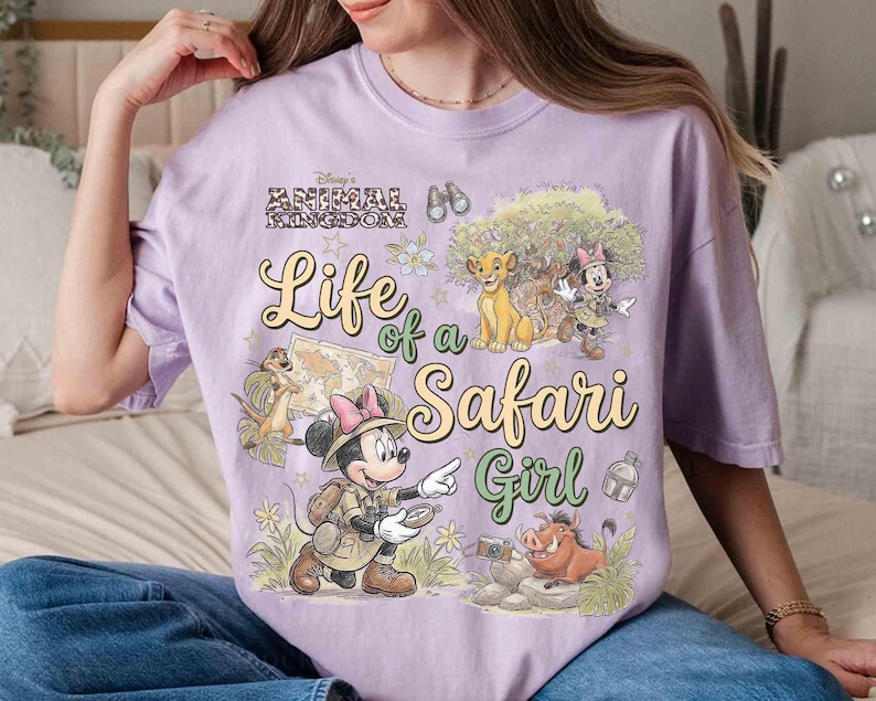 May include: A light purple t-shirt featuring a graphic design with the text "Life of a Safari Girl" and Disney's Animal Kingdom logo. The design includes illustrations of Simba, Timon, Pumbaa, and Minnie Mouse in safari attire.