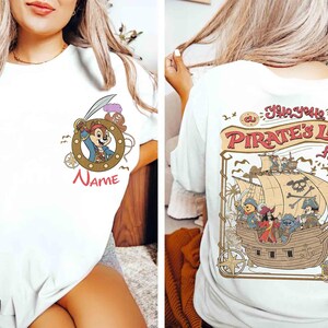 K&ouml;nnte beinhalten: Wei&szlig;es T-Shirt mit Piratenmotiv. Auf der Vorderseite befindet sich eine Cartoonfigur in einem Bullauge mit dem Wort "Name" darunter. Auf der R&uuml;ckseite ist ein Piratenschiff mit Figuren und dem Text "A Pirate's Life For Me!" abgebildet.