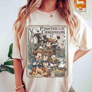 May include: Cream-colored t-shirt featuring a graphic from Disney's Pirates of the Caribbean. The design includes Mickey Mouse, Donald Duck, Goofy, and Pluto as pirates, along with a treasure chest and a pirate ship. The shirt has a crew neck and short sleeves.