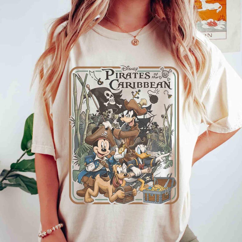 May include: Cream-colored t-shirt featuring a graphic from Disney's Pirates of the Caribbean. The design includes Mickey Mouse, Donald Duck, Goofy, and Pluto as pirates, along with a treasure chest and a pirate ship. The shirt has a crew neck and short sleeves.