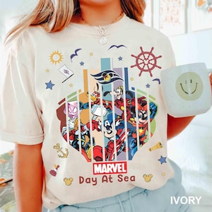 May include: Ivory-colored t-shirt featuring a colorful graphic of Marvel and Disney characters. The design includes the text "Marvel Day At Sea" and nautical elements like a ship's wheel and anchor. A white mug with a smiley face is held in the image.