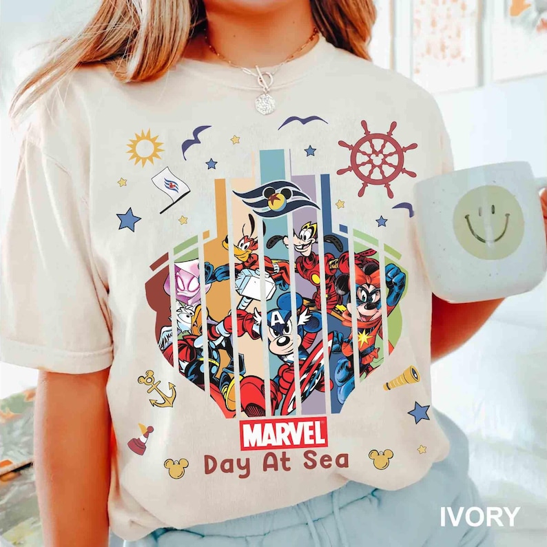 May include: Ivory-colored t-shirt featuring a colorful graphic of Marvel and Disney characters. The design includes the text "Marvel Day At Sea" and nautical elements like a ship's wheel and anchor. A white mug with a smiley face is held in the image.