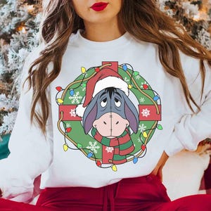 Op de afbeelding: Witte sweatshirt met een feestelijk ontwerp. Het toont Eeyore met een kerstmuts en sjaal, omlijst door een kerstkrans met lichtjes en sneeuwvlokken. Het ontwerp staat op de voorkant van de top met lange mouwen.