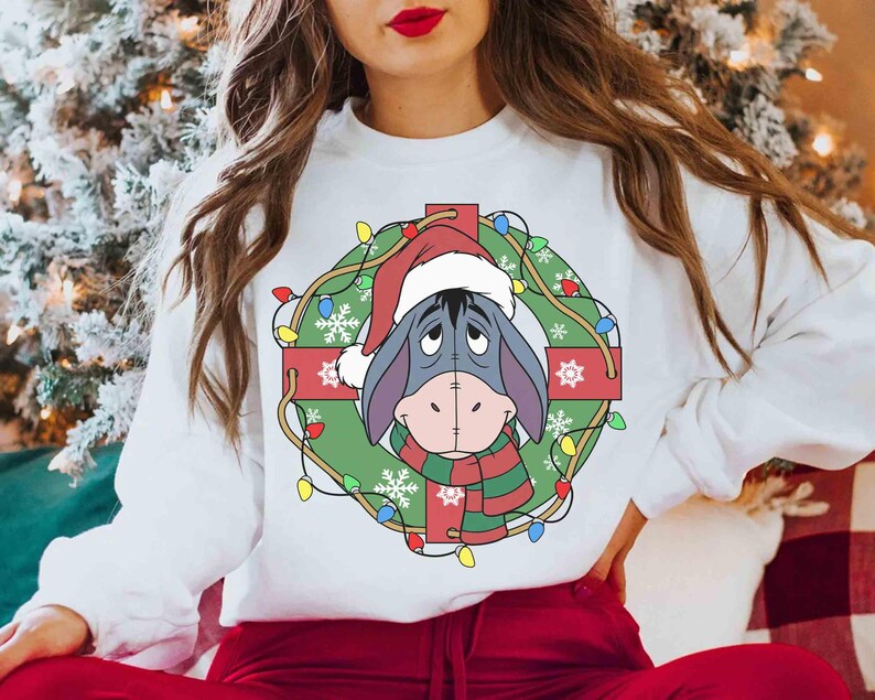 Op de afbeelding: Witte sweatshirt met een feestelijk ontwerp. Het toont Eeyore met een kerstmuts en sjaal, omlijst door een kerstkrans met lichtjes en sneeuwvlokken. Het ontwerp staat op de voorkant van de top met lange mouwen.