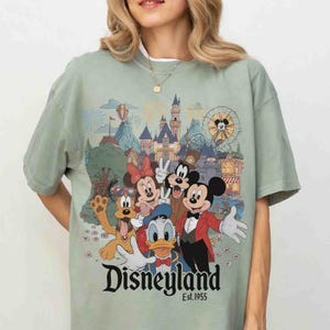 May include: Sage green t-shirt featuring a Disneyland graphic with Mickey Mouse, Minnie Mouse, Goofy, Donald Duck, and Pluto. The graphic includes a castle, Ferris wheel, and the text "Disneyland Est.1955".