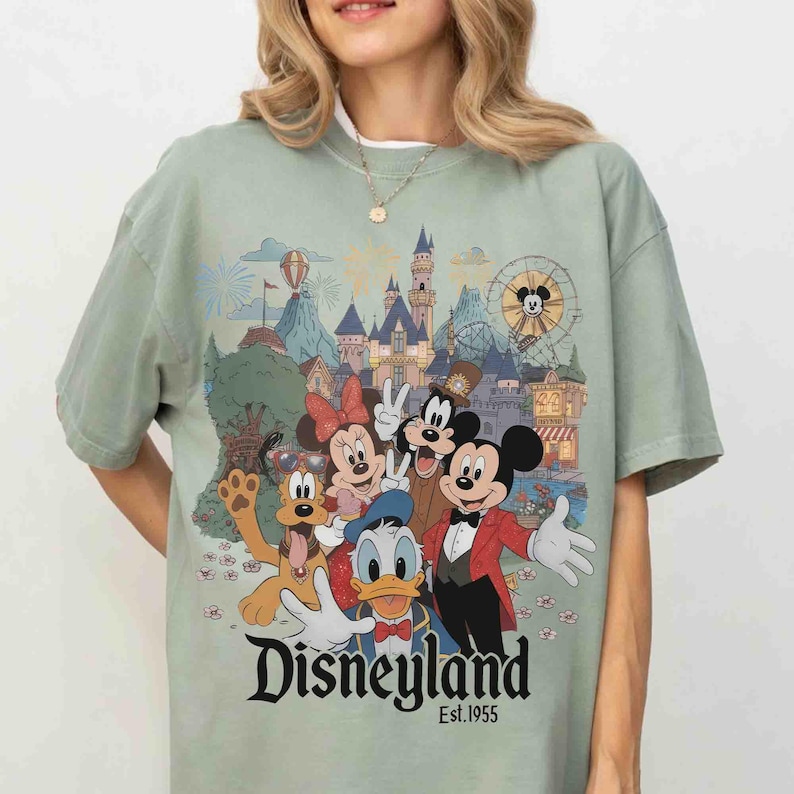 May include: Sage green t-shirt featuring a Disneyland graphic with Mickey Mouse, Minnie Mouse, Goofy, Donald Duck, and Pluto. The graphic includes a castle, Ferris wheel, and the text "Disneyland Est.1955".