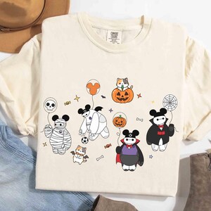 May include: Cream-colored t-shirt featuring a Halloween-themed cartoon design. The design includes characters dressed as a mummy, vampire, and other Halloween figures. The shirt has a crew neck and short sleeves. The shirt is made of a soft, comfortable material.