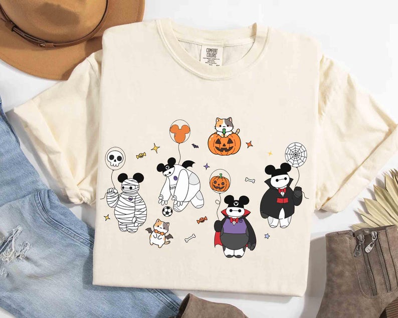 May include: Cream-colored t-shirt featuring a Halloween-themed cartoon design. The design includes characters dressed as a mummy, vampire, and other Halloween figures. The shirt has a crew neck and short sleeves. The shirt is made of a soft, comfortable material.
