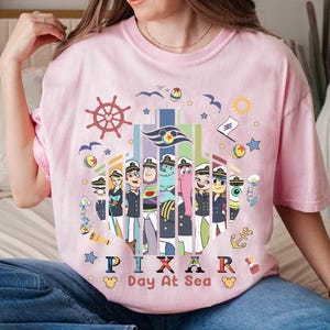 K&ouml;nnte beinhalten: Hellrosa T-Shirt mit einem farbenfrohen Grafikdesign von Pixar-Charakteren in Matrosenuniformen, mit dem Text "PIXAR Day At Sea". Das Design beinhaltet ein Steuerrad, M&ouml;wen und Sterne.