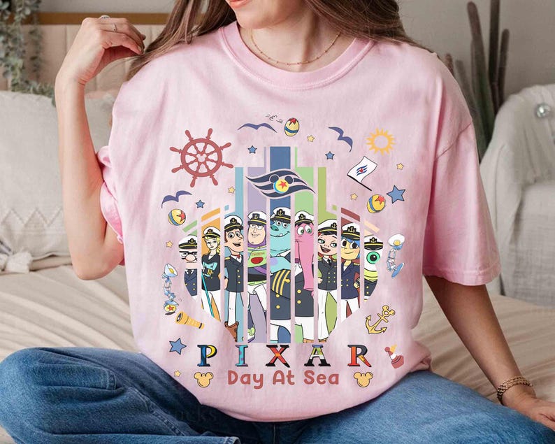 K&ouml;nnte beinhalten: Hellrosa T-Shirt mit einem farbenfrohen Grafikdesign von Pixar-Charakteren in Matrosenuniformen, mit dem Text "PIXAR Day At Sea". Das Design beinhaltet ein Steuerrad, M&ouml;wen und Sterne.