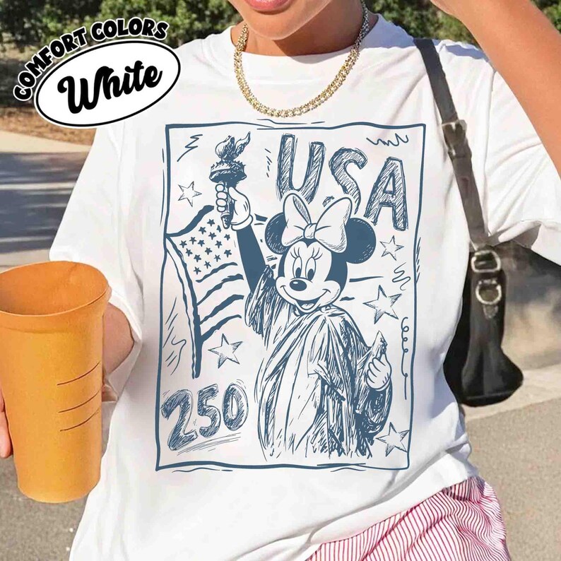 K&ouml;nnte beinhalten: Wei&szlig;es T-Shirt mit einer blauen Grafik von Minnie Mouse als Freiheitsstatue, die eine Fackel und die amerikanische Flagge h&auml;lt. Das Wort "USA" steht &uuml;ber Minnie, und die Zahl "250" steht darunter. Das Shirt hat die Worte "Comfort Colors" und "White" in einem Kreis.