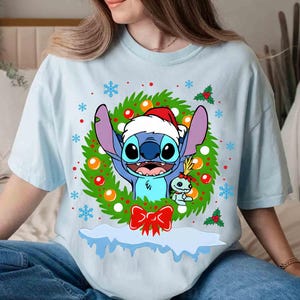 May include: Light blue t-shirt featuring a cartoon character wearing a Santa hat, surrounded by a Christmas wreath with snowflakes and a red bow. The character is blue with large ears and a friendly expression. A smaller character is also present.