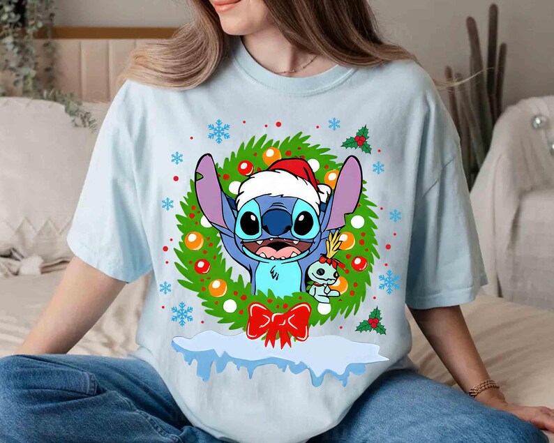 May include: Light blue t-shirt featuring a cartoon character wearing a Santa hat, surrounded by a Christmas wreath with snowflakes and a red bow. The character is blue with large ears and a friendly expression. A smaller character is also present.