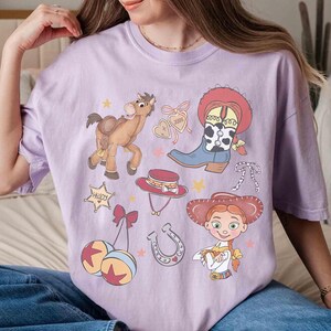 May include: A lilac t-shirt with cartoon illustrations of a cowboy theme. The design includes a cowboy boot, a horse, a sheriff's badge, a cowboy hat, a horseshoe, and a cartoon character.