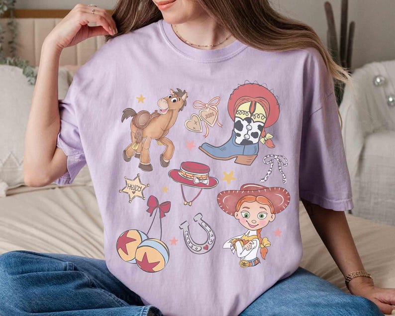 May include: A lilac t-shirt with cartoon illustrations of a cowboy theme. The design includes a cowboy boot, a horse, a sheriff's badge, a cowboy hat, a horseshoe, and a cartoon character.