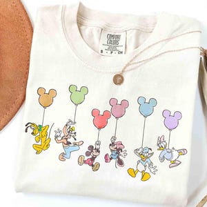 May include: Cream-colored t-shirt featuring a colorful graphic of Disney characters holding Mickey Mouse-shaped balloons. Pluto, Goofy, Mickey, Minnie, Donald, and Daisy are depicted. A gold necklace with a pendant is draped on the shirt.