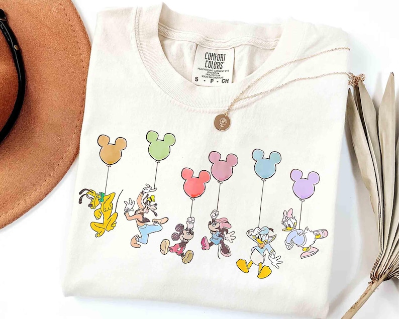 May include: Cream-colored t-shirt featuring a colorful graphic of Disney characters holding Mickey Mouse-shaped balloons. Pluto, Goofy, Mickey, Minnie, Donald, and Daisy are depicted. A gold necklace with a pendant is draped on the shirt.
