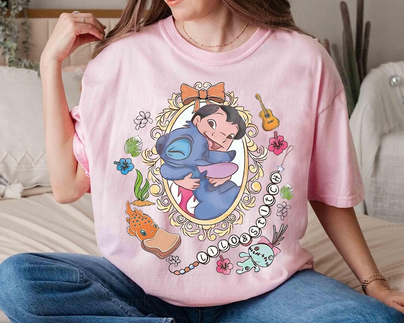 May include: Pink t-shirt featuring a cartoon graphic of Lilo and Stitch hugging inside an ornate oval frame. The design includes a guitar, flowers, and the word "Lilo & Stitch" in a chain of beads. The shirt is a crew neck.