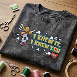 May include: A charcoal grey acid-wash t-shirt featuring an embroidered graphic of a person with long blonde hair playing a guitar. The text reads I KNEW IT, I KNEW YOU with musical notes, stars, cloud shapes, and a red heart containing the letters TS.