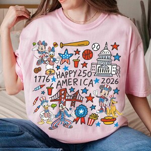 Puede incluir: Camiseta rosa claro con un dise&ntilde;o gr&aacute;fico colorido que presenta personajes de dibujos animados, estrellas y s&iacute;mbolos patri&oacute;ticos. El texto "Happy 250 America 2026" est&aacute; presente. La camiseta es de cuello redondo.