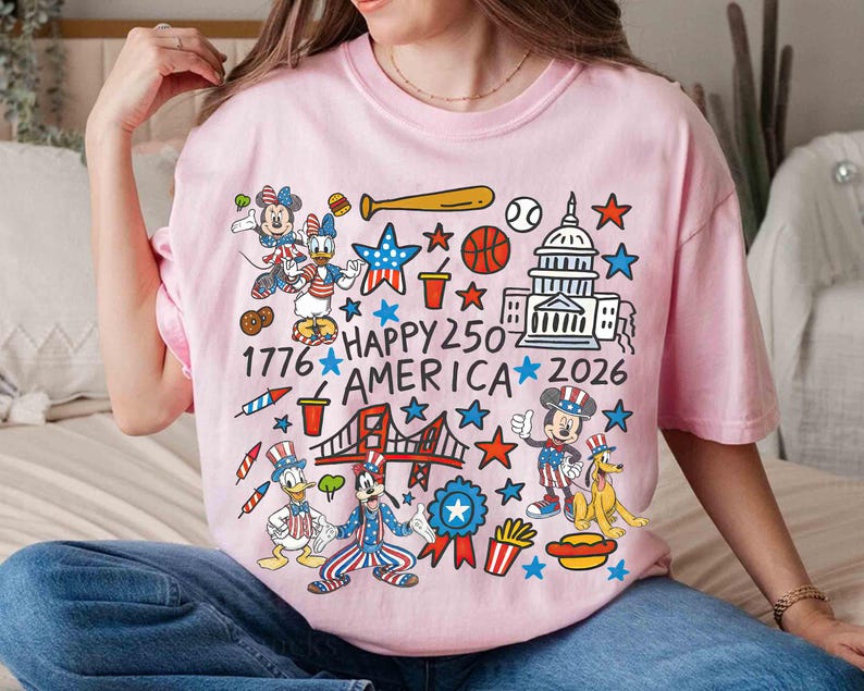 Puede incluir: Camiseta rosa claro con un dise&ntilde;o gr&aacute;fico colorido que presenta personajes de dibujos animados, estrellas y s&iacute;mbolos patri&oacute;ticos. El texto "Happy 250 America 2026" est&aacute; presente. La camiseta es de cuello redondo.