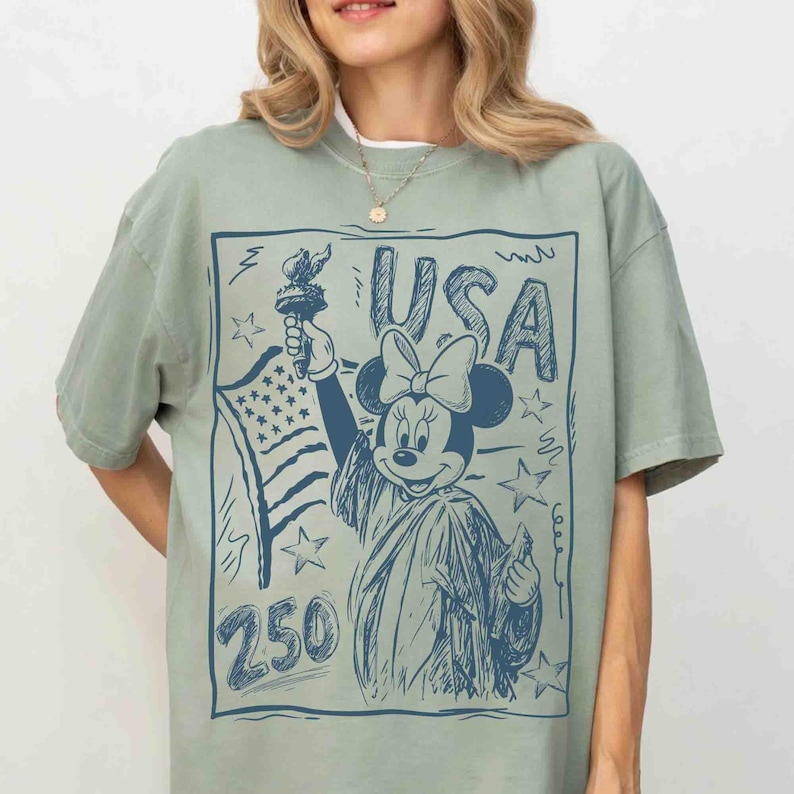 K&ouml;nnte beinhalten: Salbeigr&uuml;nes T-Shirt mit einer blauen Skizze von Minnie Mouse als Freiheitsstatue, die eine Fackel und die amerikanische Flagge h&auml;lt. Das Wort "USA" steht &uuml;ber Minnie, darunter die Zahl "250". Ein lustiges, patriotisches Design.