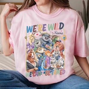 May include: Pink t-shirt featuring a colorful graphic of animated animal characters. The text "WE'RE WILD" is at the top, with "WE CAN'T BE TAMED" at the bottom. The shirt is worn by a person.