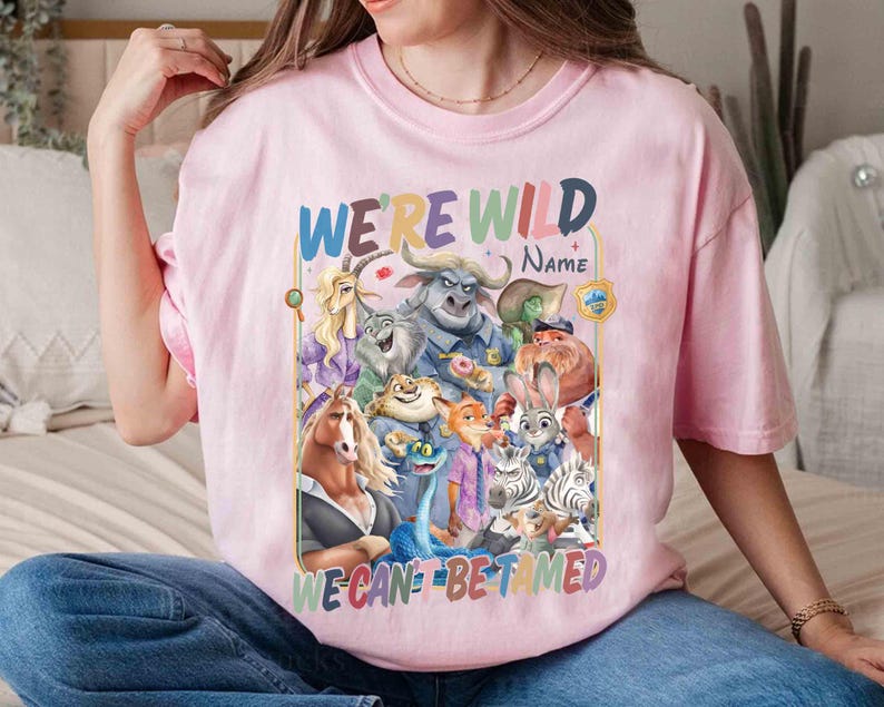 May include: Pink t-shirt featuring a colorful graphic of animated animal characters. The text "WE'RE WILD" is at the top, with "WE CAN'T BE TAMED" at the bottom. The shirt is worn by a person.