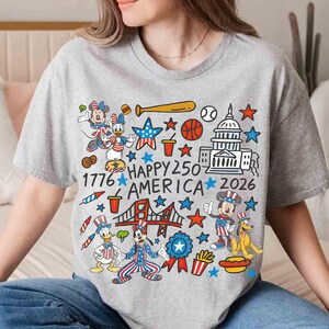 Puede incluir: Una camiseta gris con un gr&aacute;fico colorido que presenta personajes de dibujos animados, estrellas y s&iacute;mbolos patri&oacute;ticos. El texto "1776 HAPPY 250 AMERICA 2026" tambi&eacute;n est&aacute; incluido. La camiseta tiene cuello redondo y mangas cortas.