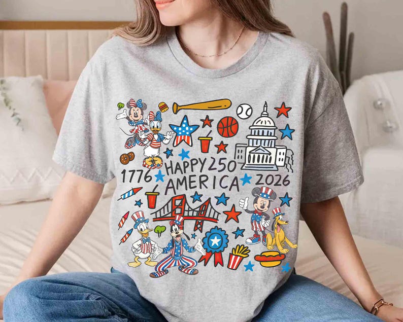 Puede incluir: Una camiseta gris con un gr&aacute;fico colorido que presenta personajes de dibujos animados, estrellas y s&iacute;mbolos patri&oacute;ticos. El texto "1776 HAPPY 250 AMERICA 2026" tambi&eacute;n est&aacute; incluido. La camiseta tiene cuello redondo y mangas cortas.