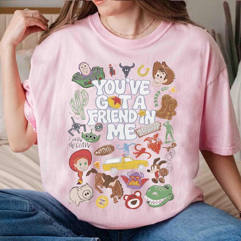 May include: Pink t-shirt featuring a colorful graphic with the text "YOU'VE GOT A FRIEND IN ME" and various cartoon character illustrations. The design includes a cactus, a cowboy hat, and a green alien.