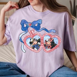 May include: A lavender t-shirt with a graphic of Mickey Mouse and Minnie Mouse in a heart-shaped locket. The locket is decorated with a blue bow and a heart.